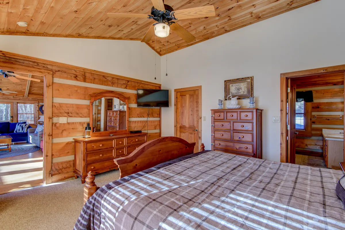 A warmly furnished bedroom with a wooden bed, comfortable linens, a dressing table and mirror, a TV mounted on the wall, and a wooden dresser. Sunlight streams in, creating a bright, welcoming space ideal for relaxation and unwinding.