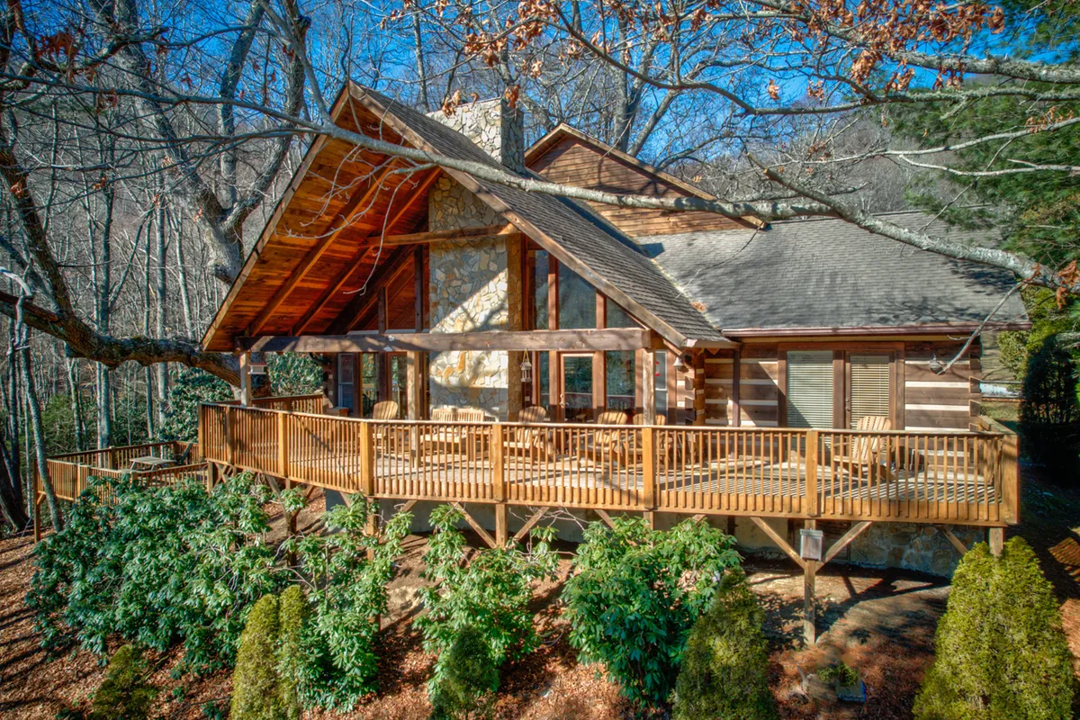 The image features the inviting exterior of a classic log cabin nestled among tall trees, with a spacious wooden deck, stone facade, and large windows that reflect the natural surroundings. The deck provides space for relaxing, gathering, and enjoying the tranquility of the forest, offering a sense of seclusion and rustic charm.