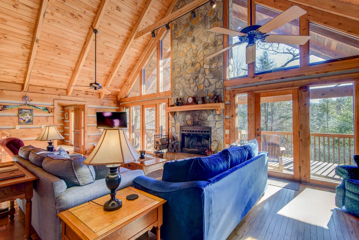 A spacious living room with a prominent stone fireplace serves as the focal point. Large windows let in ample natural light and offer soft mountain views. Comfortable seating is arranged to encourage conversation and relaxation, with visible wooden beams adding rustic charm. The overall mood is warm and inviting, ideal for gathering with family and friends.
