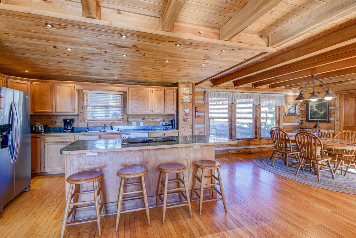 An open kitchen and dining area with a central island, wooden cabinetry, and a large dining table. Expansive windows provide plenty of daylight, making the space feel airy and lively. The layout is perfect for cooking, socializing, and sharing meals, aligning well with the communal hub described in the section.
