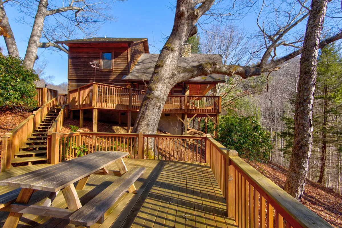 This image showcases a wooden picnic table under a large tree branch, with a staircase leading up to the house and a wooden deck visible in the background. The setting is peaceful and rustic, with the deck and yard blending into the surrounding landscape—inviting for outdoor dining, family games, and relaxed gatherings.