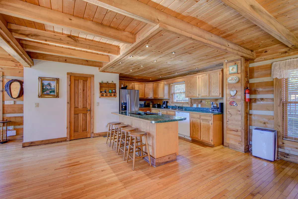 This image showcases a warm, inviting kitchen featuring a kitchen island with wooden cabinetry, dining stools, framed artwork, and exposed wooden beams. The kitchen is clearly stocked and designed for gatherings and meal preparation, with plentiful space for family or friends to join in the cooking process.