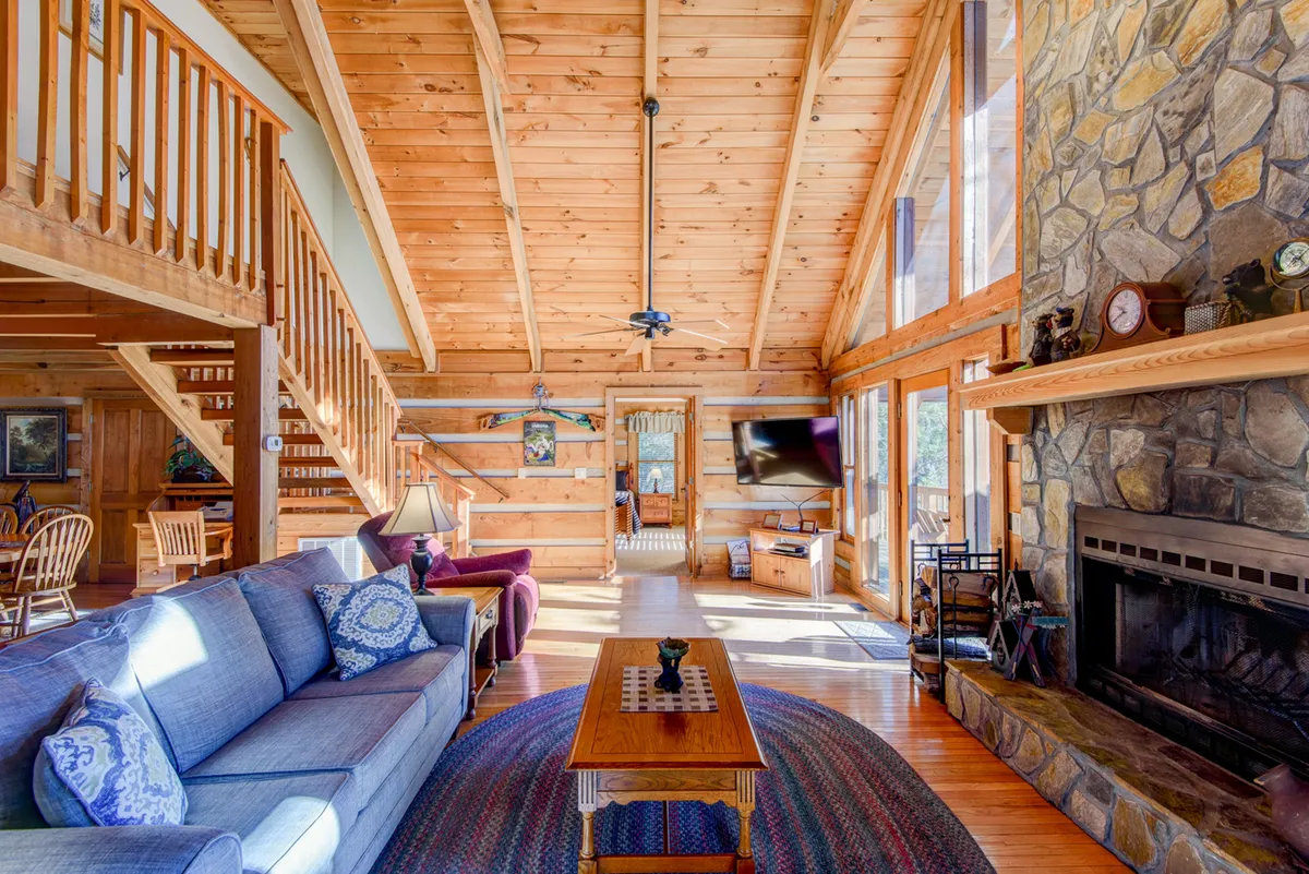 A cozy living area featuring a stone fireplace, plush seating, a wooden coffee table, and expansive windows with views of the outdoors. The wooden beams and furnishings create a harmonious, rustic atmosphere. The layout encourages group activities such as games, watching TV, and socializing by the fire.