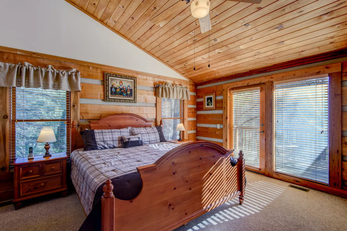 A serene bedroom featuring a wooden bed with crisp linens, framed artwork above the headboard, a cozy bedside lamp, and double doors that open to views of the outdoors. The window lets in natural light, making the space feel bright and tranquil.