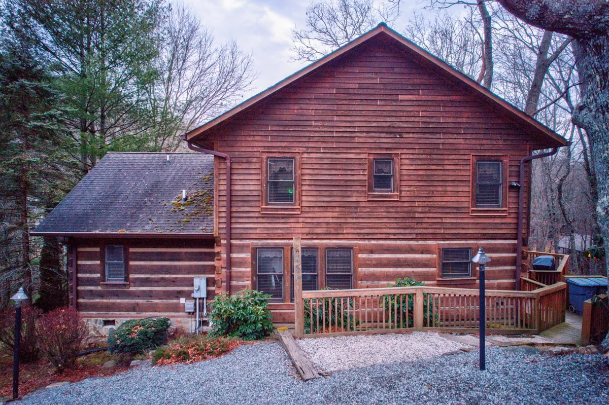 A welcoming cabin exterior with wooden siding, a spacious deck, and a beautifully landscaped garden. The gravel pathway leads up to the house, surrounded by mature trees and open yard—perfect for outdoor activities.